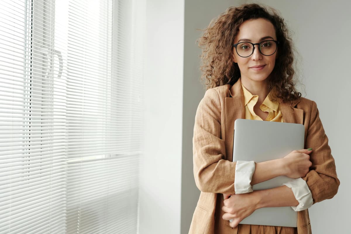 Business professional in formal attire wearing glasses, standing by a window and holding a laptop to their chest, symbolizing secure and responsible business dissolution.