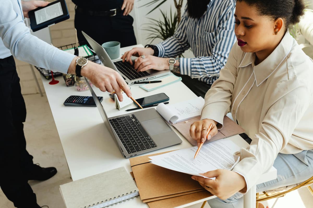 Four business professionals collaborating around a table with documents, two laptops, a phone, and a calculator, two reviewing and pointing at a document together, illustrating teamwork, document review, and strategic planning for starting a nonprofit organization.