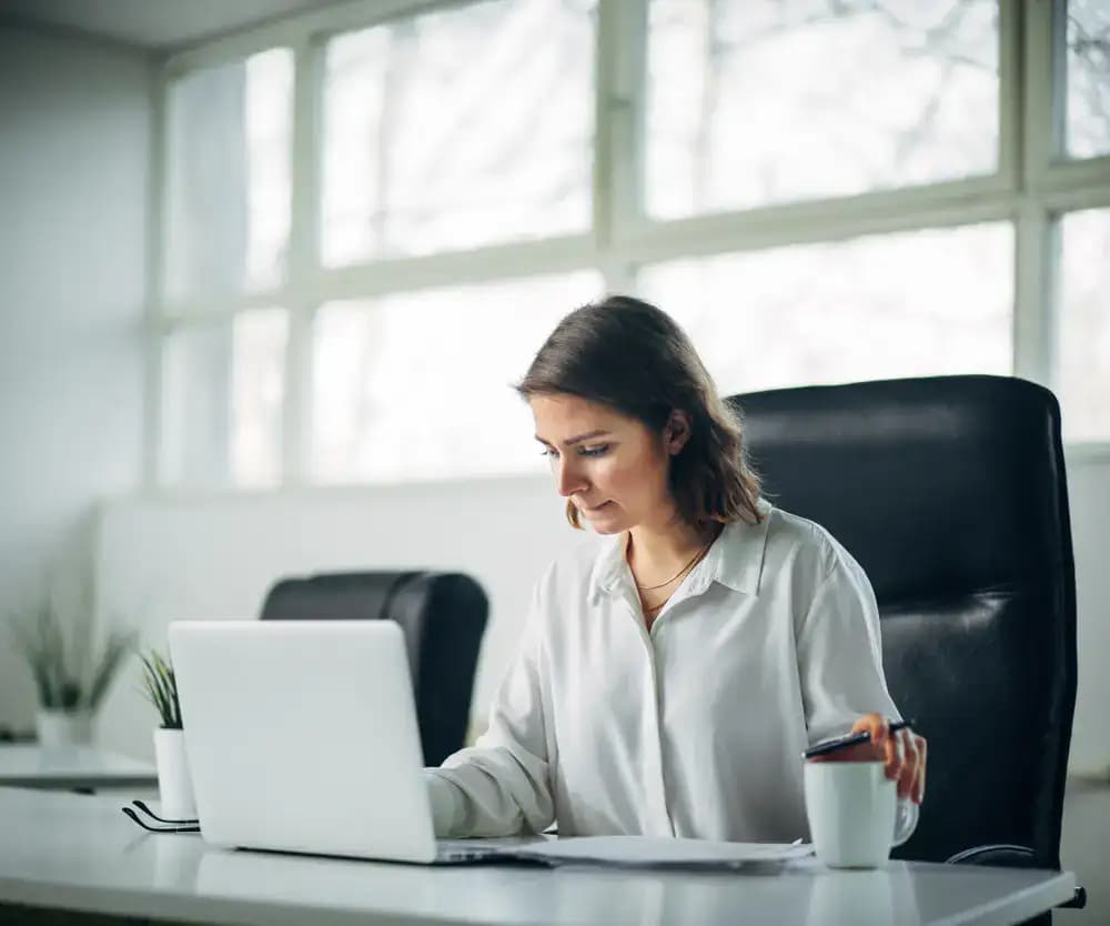 A business professional in formal attire sitting at a desk with a laptop, paperwork, and a coffee mug, focused on the screen, illustrating attentive document management and privacy protection with a registered agent service.