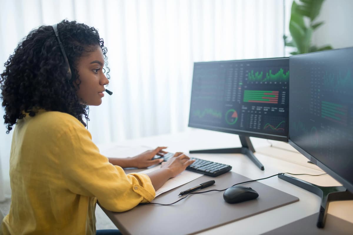 A business professional in formal attire sitting at a desk with dual monitors displaying graphs, paperwork, and a headset, demonstrating attentive document management and compliance monitoring with a registered agent service.