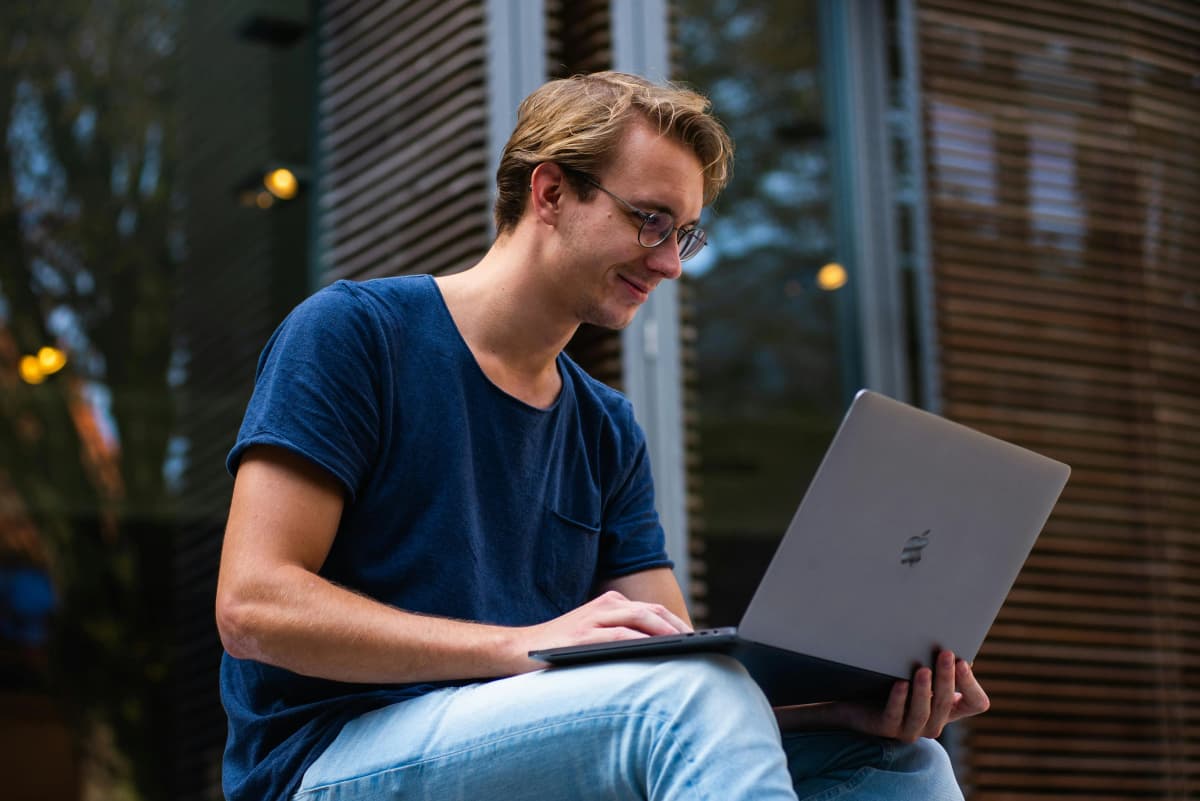 Individual business owner sitting in front of his house holding a laptop and smiling confidently, representing the ease and control of a sole proprietorship.
