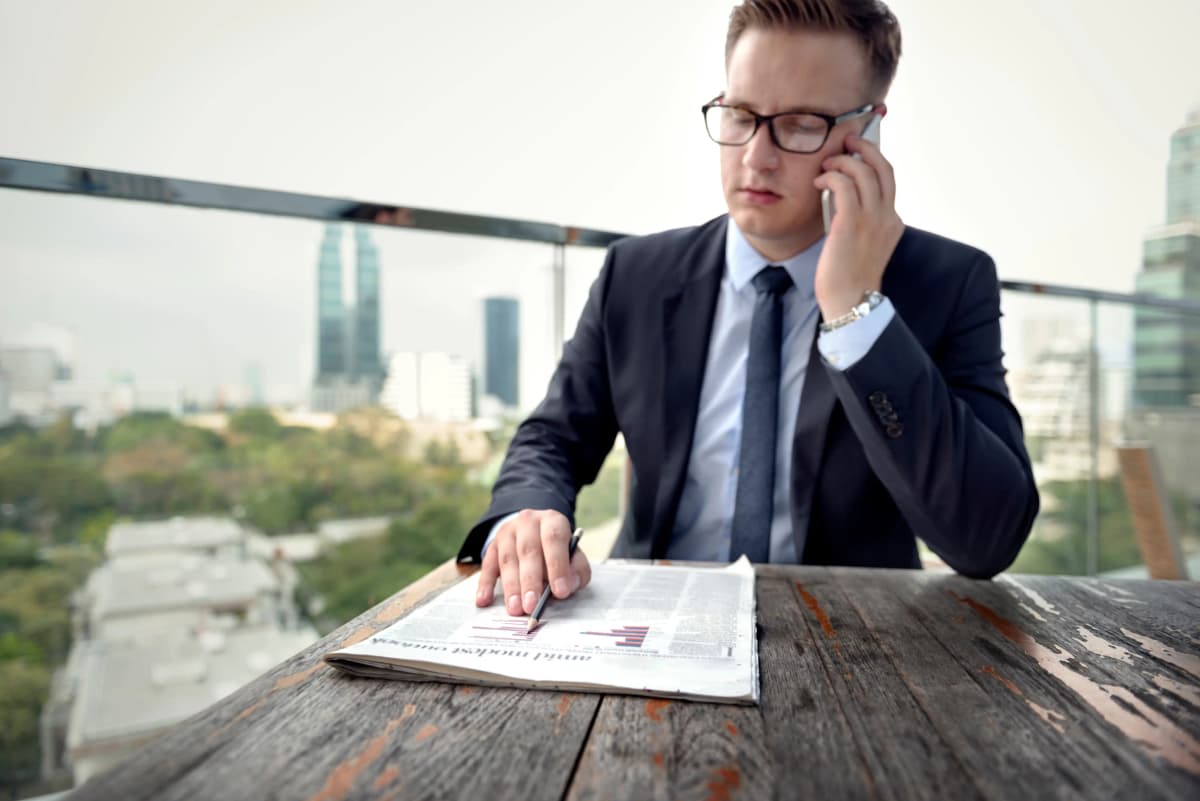 Business professional in formal attire sitting at a balcony desk with a city view, on a phone call and reviewing a paper with graphs, illustrating informed decision-making for LLC formation.