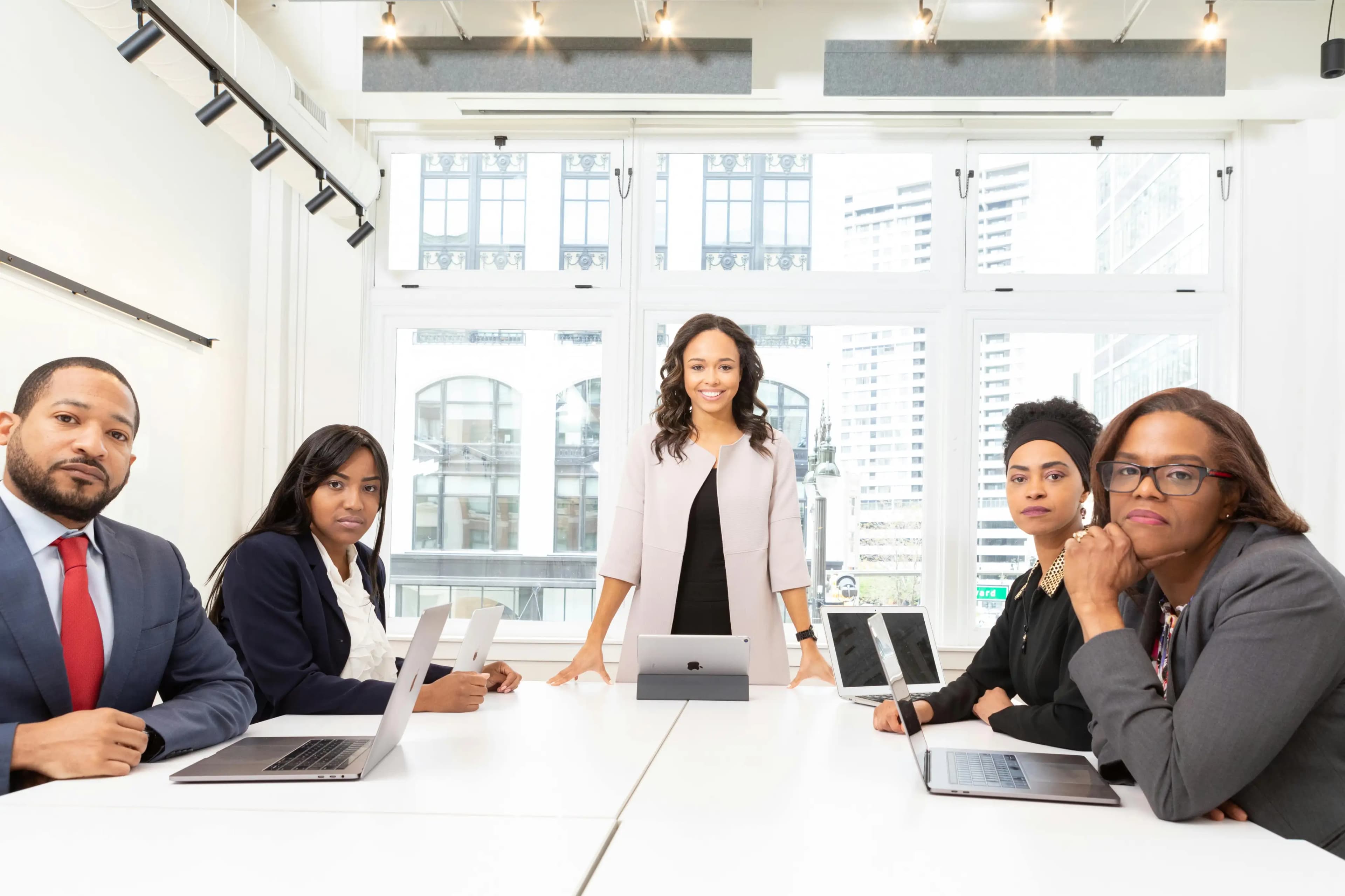 Five business professionals in formal attire gathered around a table with laptops, one standing and smiling while the others are seated, illustrating collaborative decision-making and leadership for S Corporation election and IRS compliance, with a city view in the background.